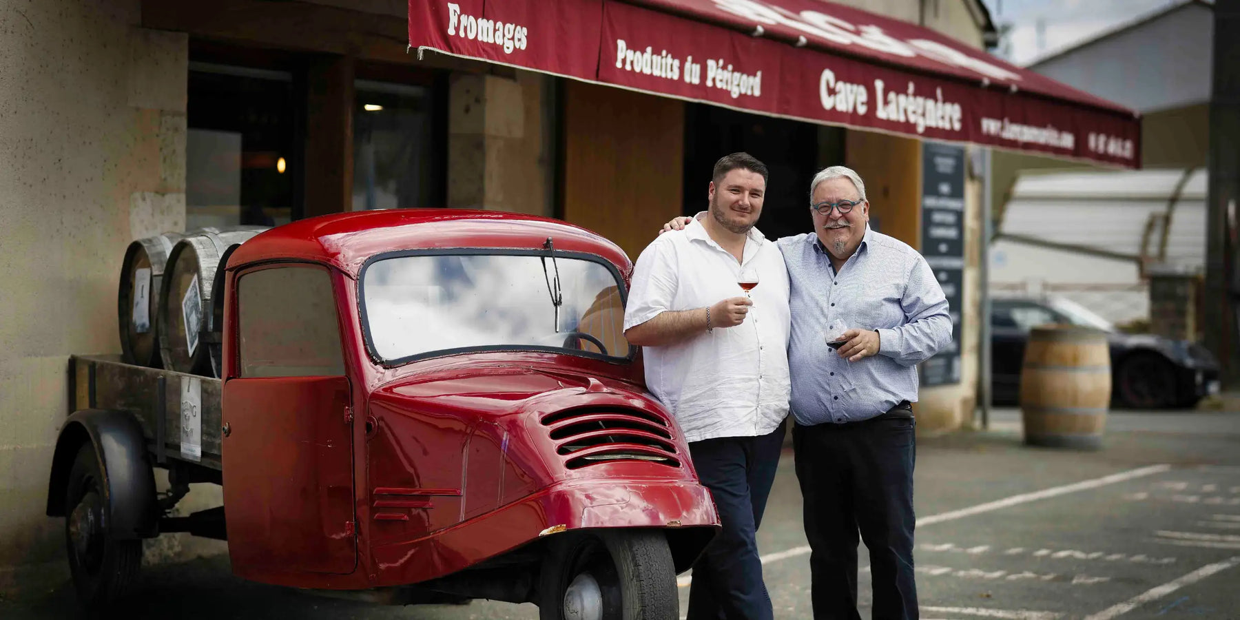 Michel et Grégoire Baraton devant la Cave Larégnère, un verre à la main, avec le triporteur rouge de la maison